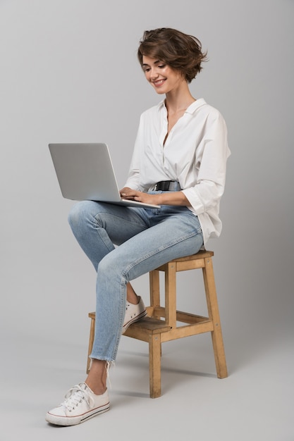 Het jonge bedrijfsvrouw stellen geïsoleerd over grijze muurzitting op kruk die laptop computer met behulp van