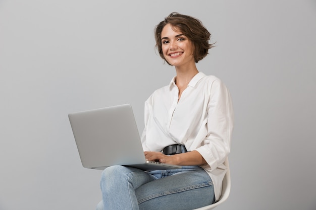 Het jonge bedrijfsvrouw stellen geïsoleerd over grijze muurzitting op kruk die laptop computer met behulp van