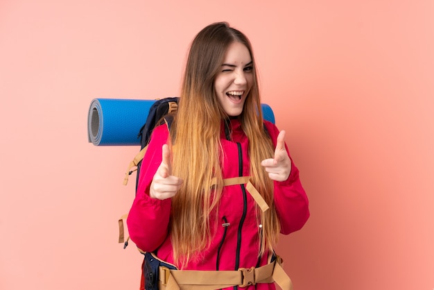 Het jonge bergbeklimmermeisje met een grote rugzak op roze muur richt vinger op u