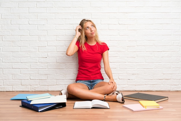 Het jonge blonde studentenmeisje met vele boeken op de vloer die twijfels hebben en verwart gezichtsuitdrukking