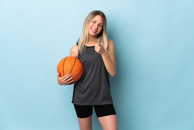 Het jonge blonde vrouw speelbasketbal dat op blauwe muur wordt geïsoleerd, wijst vinger naar u