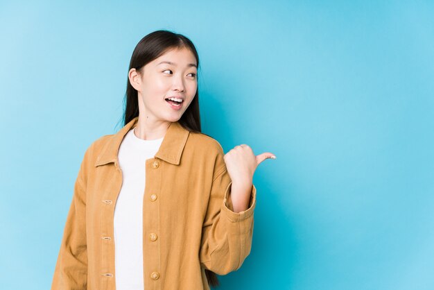 Het jonge Chinese vrouw stellen in een blauwe muur geïsoleerde punten met weg duim, lachend en onbezorgd.