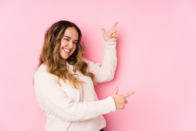 Het jonge curvy vrouw stellen in een roze geïsoleerde muur het richten met wijsvingers aan een exemplaarruimte, die opwinding en wens uitdrukken.