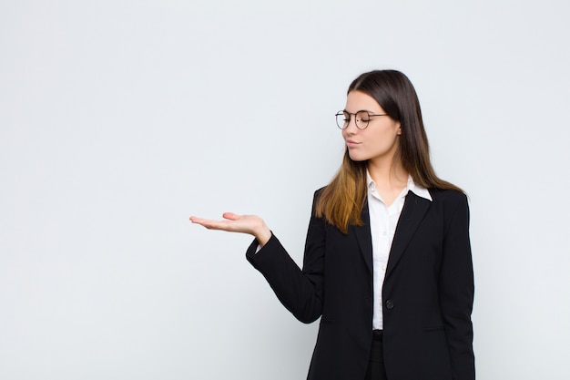 Het jonge gelukkig voelen en onderneemster die terloops glimlachen, kijkend aan een voorwerp of een concept hield op de hand aan de kant tegen witte muur