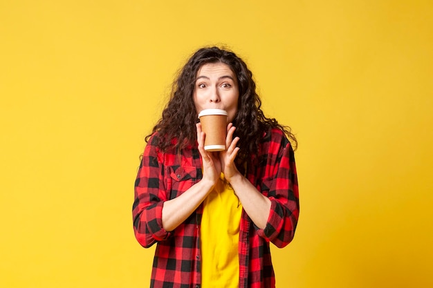 Het jonge geschokte donkerbruine meisje in een plaidoverhemd drinkt koffie en toont verrassing op een gele achtergrond