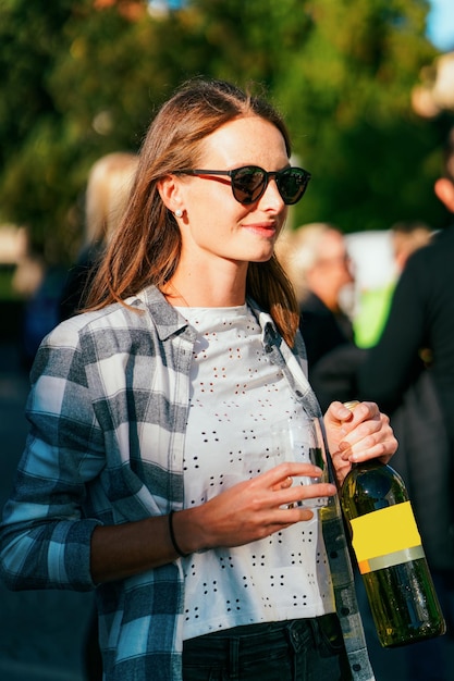 Het jonge glimlachende meisje dat zonnebril draagt houdt wijnfles en glas vast. Vrouw casual stijl. Vrouwelijke persoon heeft plezier. Fijne vakantieviering. Buiten levensstijl portret. Volwassen mooi gezicht. straat evenement