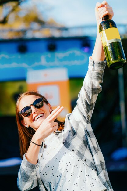 Het jonge glimlachende meisje dat zonnebril draagt houdt wijnfles. Vrouw casual stijl. Vrouwelijke persoon die plezier heeft. Fijne vakantieviering. Buiten levensstijl portret. Volwassen mooi gezicht. straat evenement