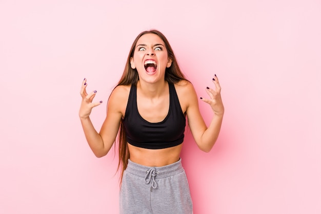 Het jonge Kaukasische geschiktheidsvrouw stellen in een roze muur die met woede gilt.