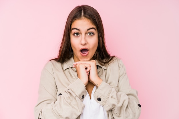 Het jonge Kaukasische vrouw stellen in een roze muur die voor geluk bidden, verbaasde en mond openen die aan voorzijde kijken.