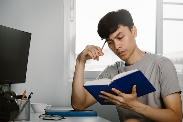 Het jonge knappe aziatische boek van de mensenlezing en het denken gebaar bij het werkbureau in vrije tijd van thuis het werken, kennis en het leren concept