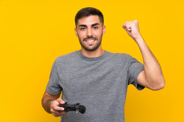Het jonge knappe mens spelen met een videospelletjecontrolemechanisme over geïsoleerde gele muur die een overwinning vieren