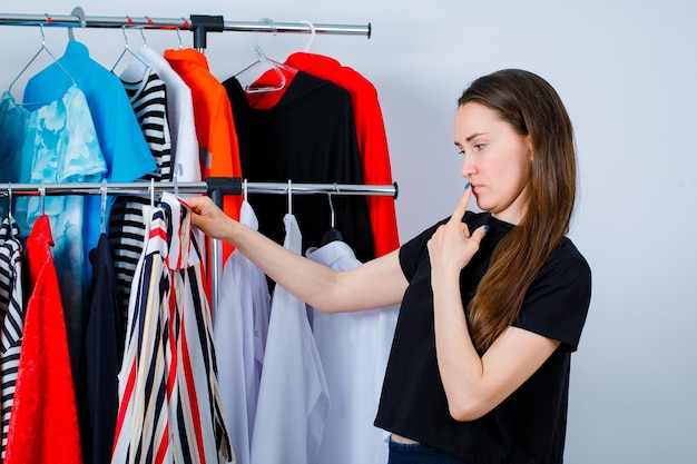 Het jonge meisje denkt door kleurrijke blouse op klerenachtergrond te houden