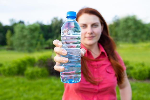 Het jonge meisje in heldere de zomerkleren houdt een plastic fles water op de achtergrond van het park