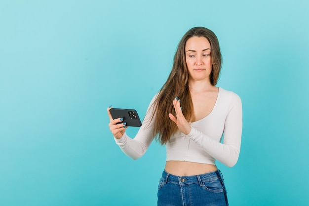 Het jonge meisje kijkt naar het smartphonescherm door een stopgebaar op een blauwe achtergrond te tonen