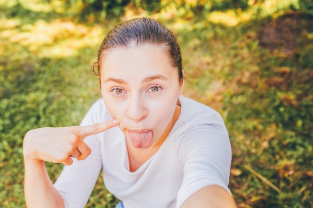 Het jonge meisje neemt selfie uit handen met telefoon die tong en grappige gezichtszitting op park of tuin tonen. Portret van jonge aantrekkelijke vrouw die selfie foto op smartphone in de zomerdag.