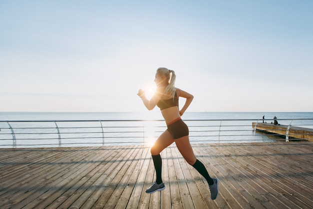 Het jonge mooie atletische meisje met lang blond haar in zwarte kleren die bij zonsopgang boven de zee loopt