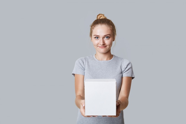 Het jonge mooie blondewijfje met haarbroodje houdt wit vakje in handen, met exemplaarruimte voor uw tekst, die over grijze achtergrond wordt geïsoleerd. Aantrekkelijke vrouw toont doos, heeft een aantrekkelijk uiterlijk