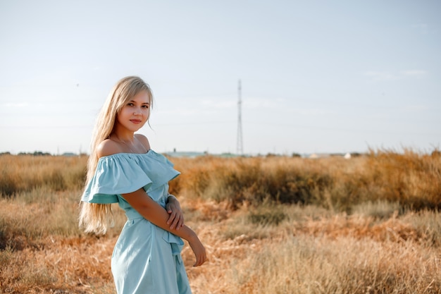 Het jonge mooie Kaukasische blondemeisje in lichtblauwe kleding bevindt zich op het gebied met het zon-geschroeide gras