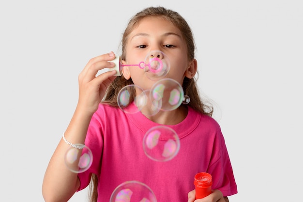 Het jonge mooie meisje draagt roze T-shirt blazende zeepbels tegen een grijze achtergrond
