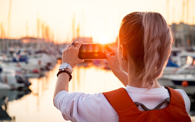 Het jonge mooie meisje neemt een foto van een baai met jachten bij zonsondergang