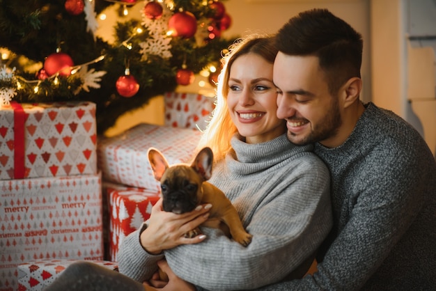 Het jonge mooie paar speelt met hun hond in feestelijke nieuwjaarswoonkamer vlak voor Kerstmis