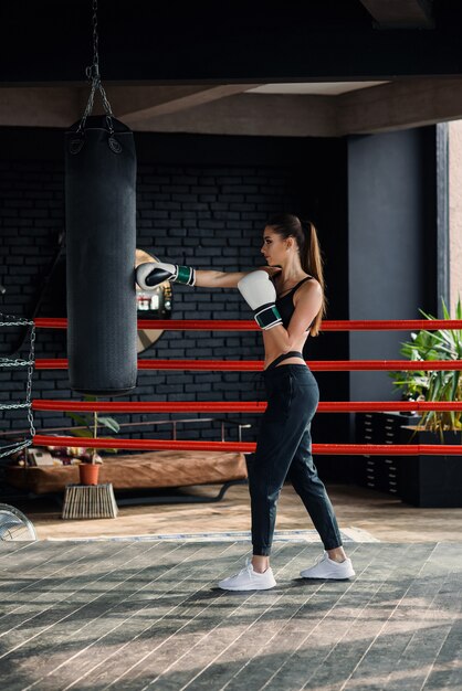 Het jonge mooie sportenmeisje in sportkleding en bokshandschoenen raakt een bokszak in moderne zwarte gymnastiek.