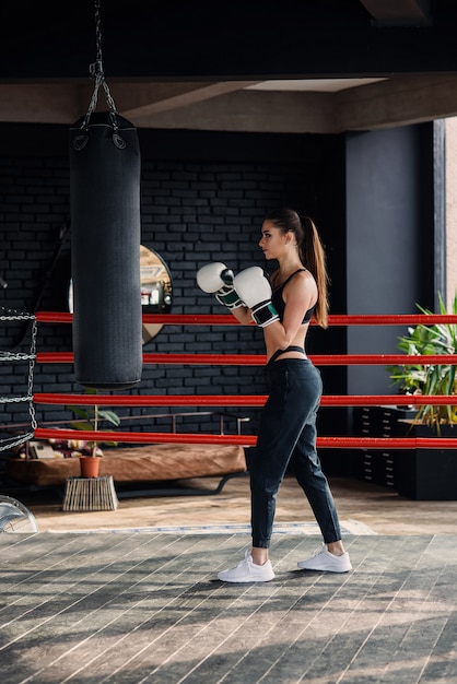 Het jonge mooie sportenmeisje in sportkleding en bokshandschoenen raakt een bokszak in moderne zwarte gymnastiek.