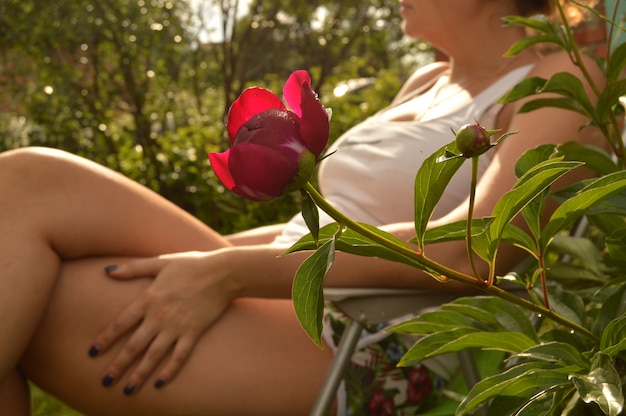Het jonge vrouw ontspannen als zitkamervoorzitter in haar tuin, dichtbij een mooie pioenbloem