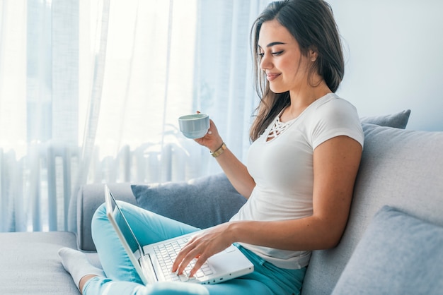 Het jonge vrouw ontspannen thuis op de laag, heeft zij een koffie en gebruikt laptop