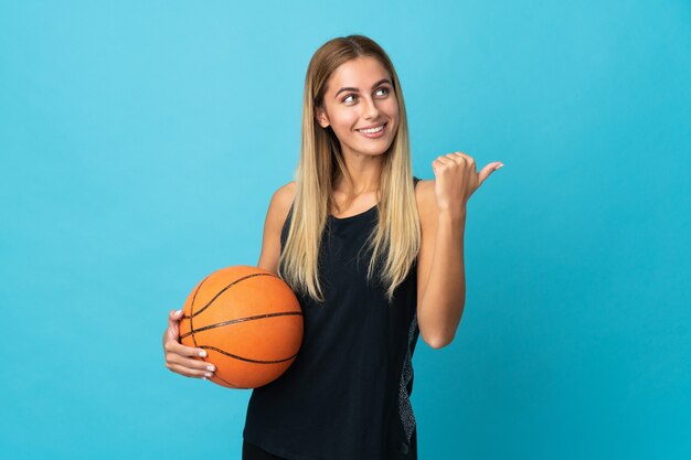 Het jonge vrouw speelbasketbal dat op witte achtergrond wordt geïsoleerd die naar de kant wijst om een product te presenteren