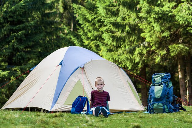 Het kamperen van de vakantie. Gelukkige jonge jongenszitting voor een tent dichtbij rugzakken die rust nemen na wandeling in het bos.