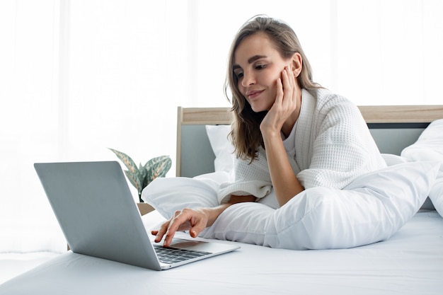 Het Kaukasische vrouw in pyjama sociaal spelen in laptop op wit bed.