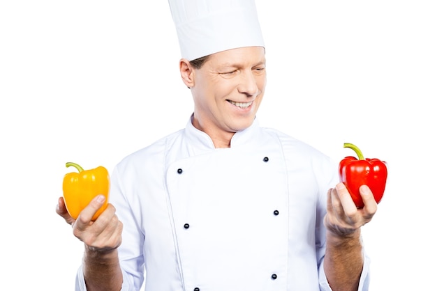 Het kiezen van de juiste peper voor zijn maaltijd. Vrolijke volwassen chef-kok in wit uniform die kleurrijke paprika's vasthoudt en naar de rode kijkt terwijl hij tegen een witte achtergrond staat