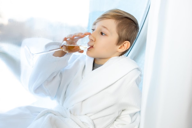 Het kind houdt een glas alcoholvrije kinderchampagne voor zich en drinkt het zittend bij het raam