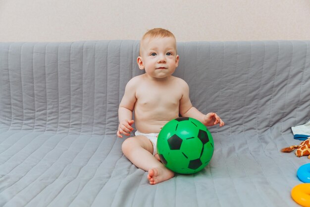 Het kind houdt een voetbal vast terwijl hij blootsvoets op de bank zit en wegkijkt Schattige baby speelt thuis op de bank Het concept van vakantie, weekends en jeugd