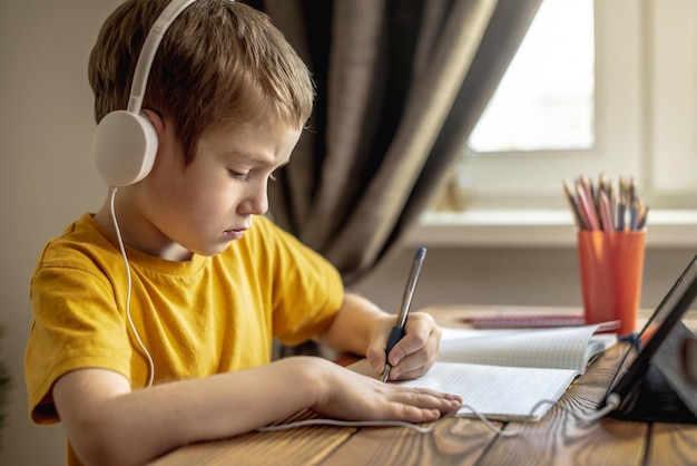 Het kind is een jongen in een gele T-shirt met een koptelefoon die huiswerk maakt op een tablet Concept van onderwijs op afstand