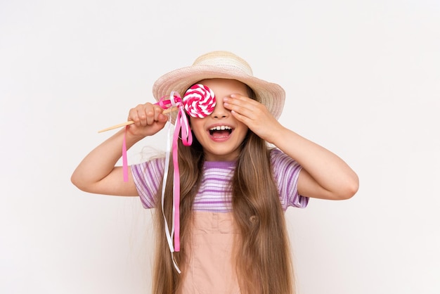 Het kind sluit één oog met een karamellolly een klein meisje met lang haar in een zomerjurk en een strohoed op een witte geïsoleerde achtergrond