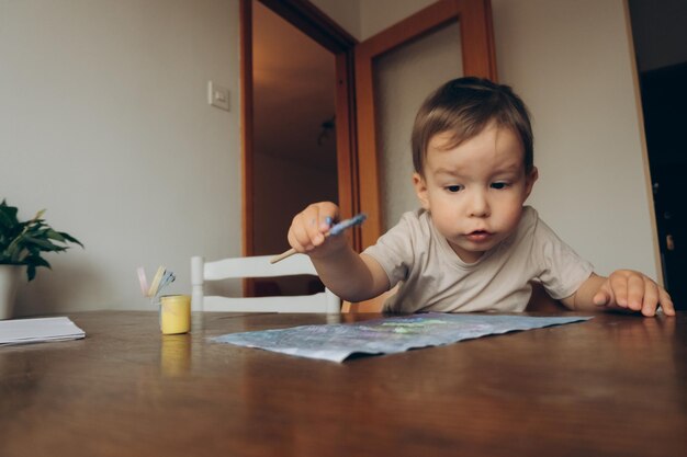 het kind tekent met een penseel en veelkleurige plakkaatverf terwijl hij aan tafel zit te tekenen naar zijn moeder