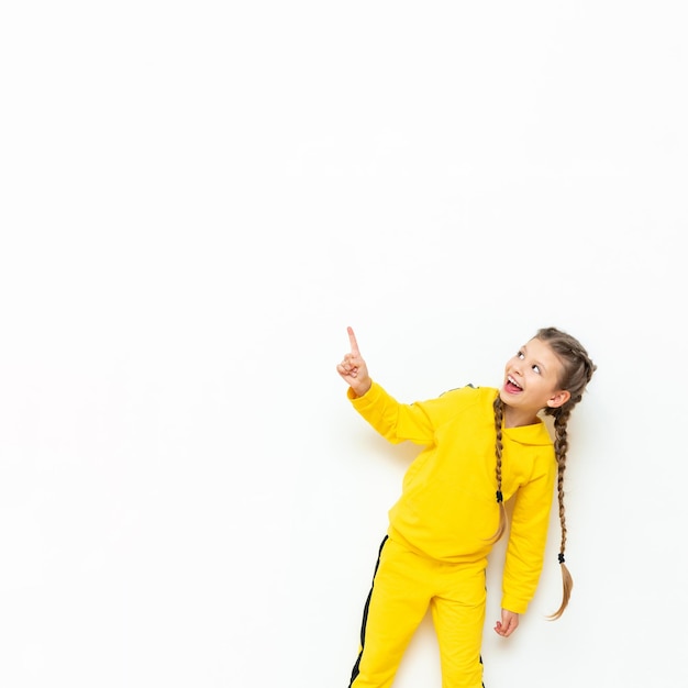 Het kind wijst naar uw advertentie Een klein meisje met twee staartjes in een geel sportpak op een witte geïsoleerde achtergrond