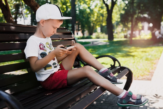 Het kind zit in het park op een bank met een gadget. Kinderen gebruiken gadgets. Portret van een mooie jongen in de ondergaande zon. Een jongen speelt een spel op een mobiele telefoon.