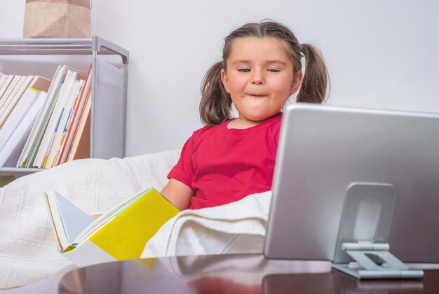 Het kind zit met een boek en wordt afgeleid door de tablet op de tafel bij de stoel