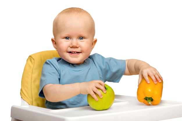 Het kind zit op een hoge stoel met een groene appel en een gele paprika in zijn hand glimlachend