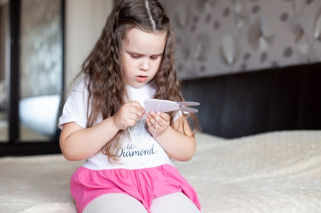 Het kindmeisje naait een stuk speelgoed gemaakt van vilt zittend op het bed op een beige deken; handwerk