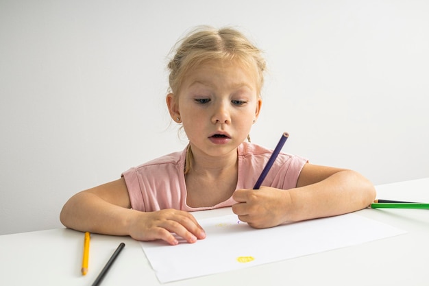 Het kindmeisje tekent met kleurpotloden zittend aan een witte tafel op een witte achtergrond