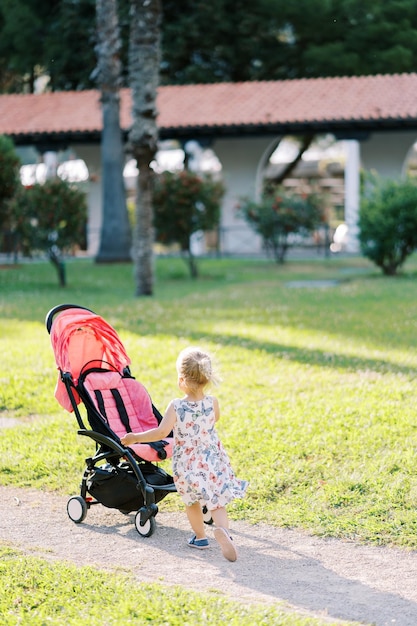 Foto het kleine meisje gaat naar een kinderwagen op een pad in een zonnig park achter uitzicht