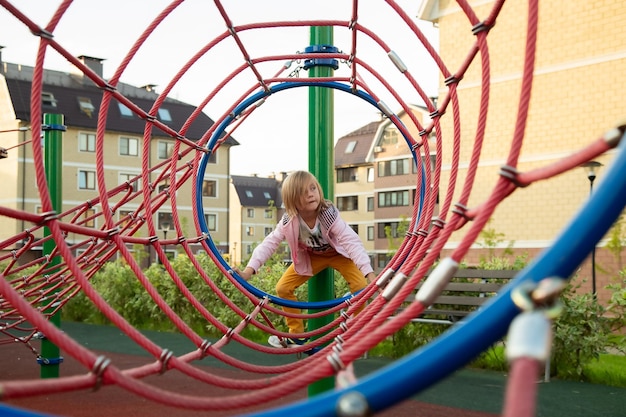 Het kleine meisje klimt touwen op de speelplaats tegen de achtergrond van huizen in de zomer