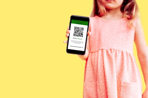 Het kleine meisje met de mobiele telefoon een model voor digitaal vaccinatiecertificaat in de ene hand en paspoort, masker en ticket in een andere op gele achtergrond