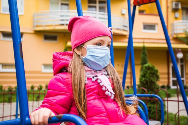 Het kleine verdrietige meisje draagt een beschermend masker zittend op een schommel Het eenzame meisje in een stadsspeeltuin in de herfst