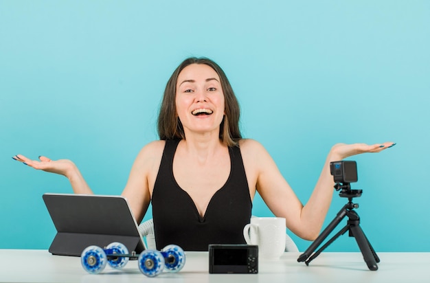 Het lachende bloggermeisje kijkt naar de camera door haar handen wijd te openen op een blauwe achtergrond