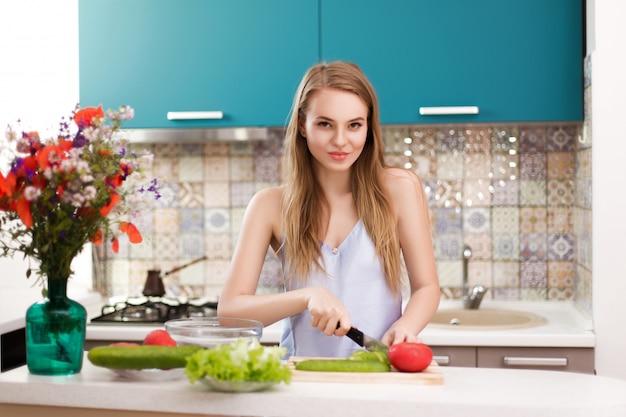 Het leuke blonde snijdt groenten voor salade op een achtergrond van blauwe keuken